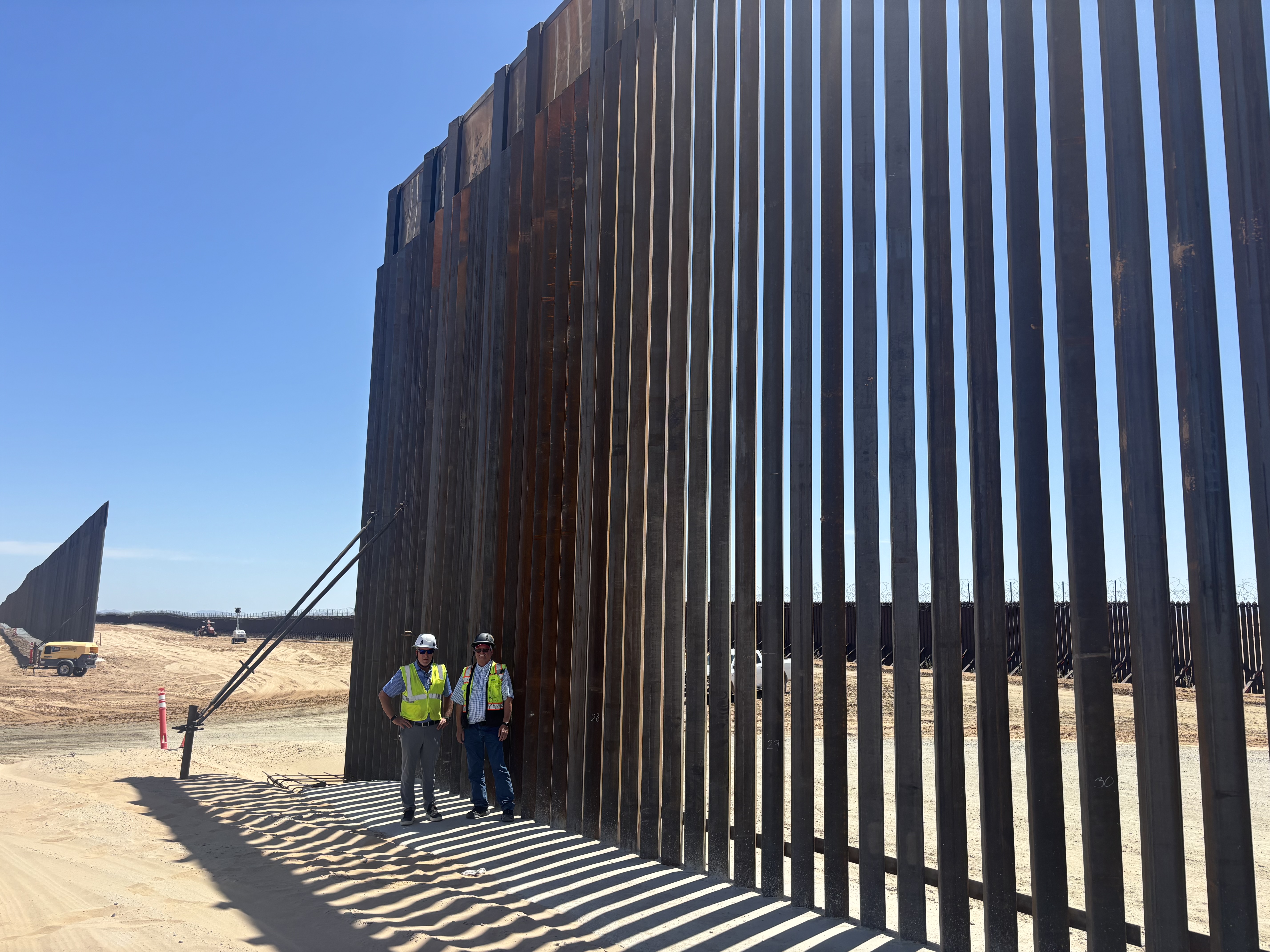 Field-visit participants standing beside the towering steel border fence in California